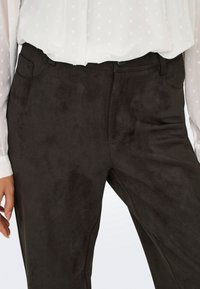Gros plan sur un pantalon en daim noir avec poches avant, porté avec un chemisier blanc à manches longues présentant un motif de cœur subtil.