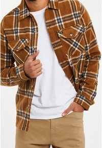 Braunes karierte Flanellhemd mit weißen und blauen Akzenten, zwei Brusttaschen und Knopfverschluss. Getragen über einem weißen T-Shirt und beigefarbenen Hosen.