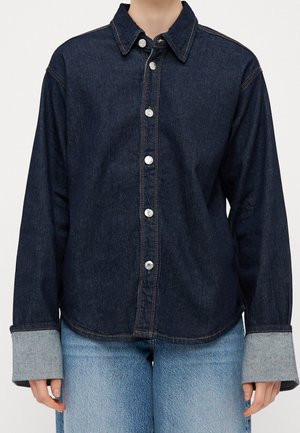 Persona que lleva una camisa vaquera oscura abotonada con puños grises doblados y vaqueros de mezclilla azul claro sobre un fondo liso.