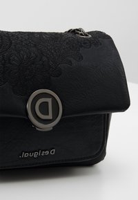 Černá kabelka z umělé kůže s krajkovým vzorem, stříbrné kování a kulatým zapínáním s logem 'D'; hladký povrch a obdélníkový tvar.