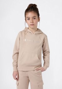 Jong meisje met bruin haar in een knot, gekleed in een beige Champion hoodie en bijpassende cargobroek, staand met één hand in de zak.