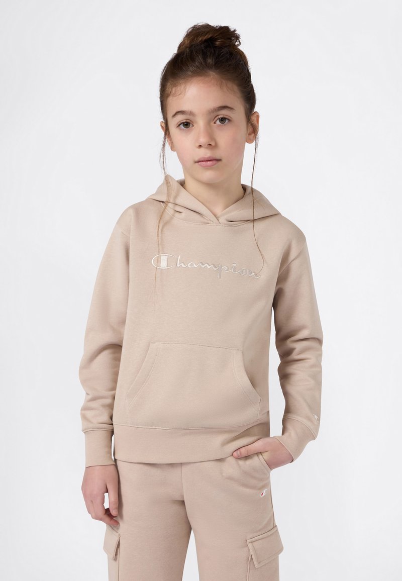 Jong meisje met bruin haar in een knot, gekleed in een beige Champion hoodie en bijpassende cargobroek, staand met één hand in de zak.