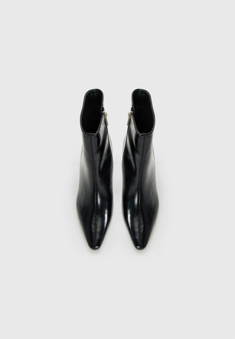 Un paio di stivali alla caviglia neri in pelle con punta affilata e cerniere laterali, esposti dall'alto su uno sfondo chiaro e semplice.