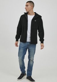 Black fleece zip-up jacket with hood, front zip pocket, and ribbed cuffs, paired with light blue jeans and black sneakers.