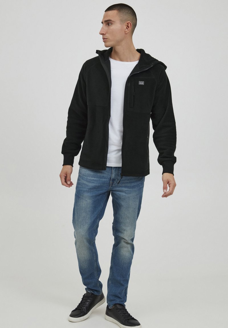 Black fleece zip-up jacket with hood, front zip pocket, and ribbed cuffs, paired with light blue jeans and black sneakers.