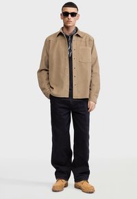 Taninuppudega särk rinnataskuga, kantud tume musta T-särgi peal, koos mustade lahtiste pükste ja tan värvi saapadega. Lisatud tumedad päikeseprillid.