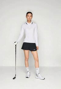 Moteriška golfo apranga, kurioje yra balta ilgomis rankovėmis polo marškiniai su juodais akcentais, juoda plisuota sijonė, baltos golfo bateliai ir golfo lazda.