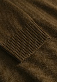 Brown knitted sweater fabric with ribbed cuff texture. Close-up detail showing soft, warm texture and subtle variation in stitching.