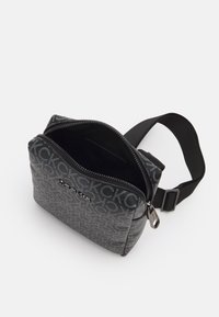 Šedá látková crossbody taška s vzorovaným logem, zipovým uzávěrem a nastavitelným černým popruhem. Interiér je podšitý černou látkou.