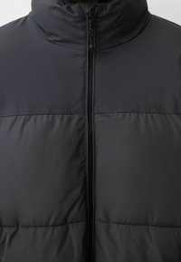 Gros plan d'une veste matelassée noire avec un col montant et une fermeture éclair centrale noire entièrement fermée.