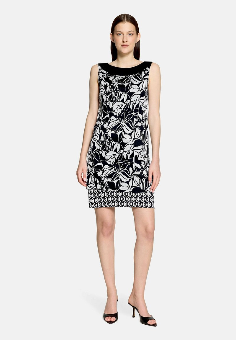 Femme debout dans une robe sans manches noir et blanc à motifs floraux et géométriques, portant des talons hauts noirs ouverts à l’avant, sur un fond uni.