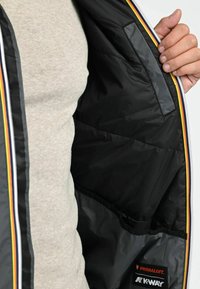 Zwarte K-Way jas met een Primaloft interieur, een ritssluiting aan de voorkant en kleurrijke gestreepte accenten langs de randen. Soepele, matte afwerking.