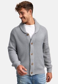 Grauer gestrickter Cardigan mit einem Schalkragen, fünf Holzknöpfen, gerippten Bündchen und einem strukturierten Muster.Über einem weißen T-Shirt getragen.