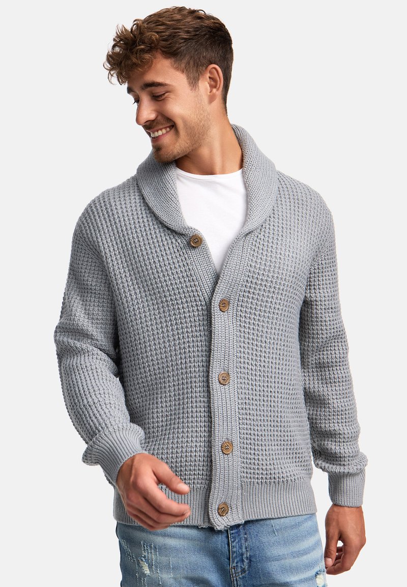 Grauer gestrickter Cardigan mit einem Schalkragen, fünf Holzknöpfen, gerippten Bündchen und einem strukturierten Muster.Über einem weißen T-Shirt getragen.