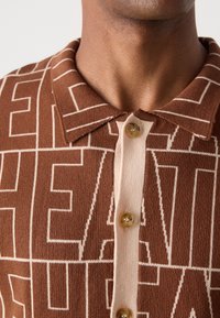 Brown knit polo shirt featuring a patterned design with cream accents, a beige button placket, and two round buttons at the collar.