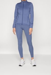Blå idrottsjacka med dragkedja och sidofickor, i kombination med matchande leggings. Texturen verkar slät och elastisk.