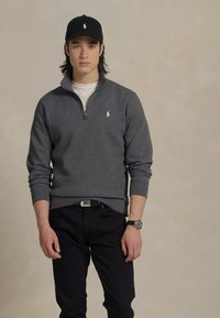 Felpa grigia con zip e logo bianco, indossata sopra una t-shirt bianca. Pantaloni neri e berretta nera con logo coordinato. Orologio da polso argento.