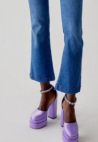 Tacchi a piattaforma viola con una finitura lucida, caratterizzati da un spesso tacco a blocco e cinghie alla caviglia decorate con piccoli gemme trasparenti, indossati con jeans blu.