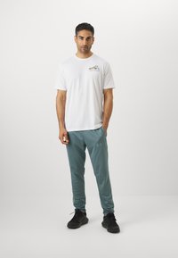 Hvid bomulds t-shirt med et grafisk design, kombineret med teal træningsbukser lavet af letvægtsmateriale. Derudover sorte sneakers.