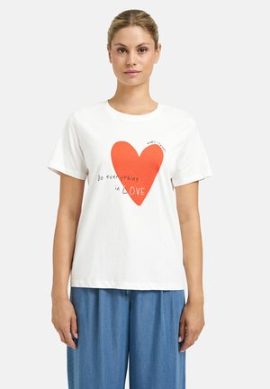 Woman wearing white T-shirt with large red heart and text "Do everything in love," paired with loose blue pants, standing against plain background.