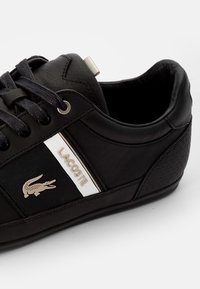 Baskets en cuir noir avec détails texturés, étiquette logo métallique et bande blanche. Elles présentent des lacets noirs et une forme à bout rond.