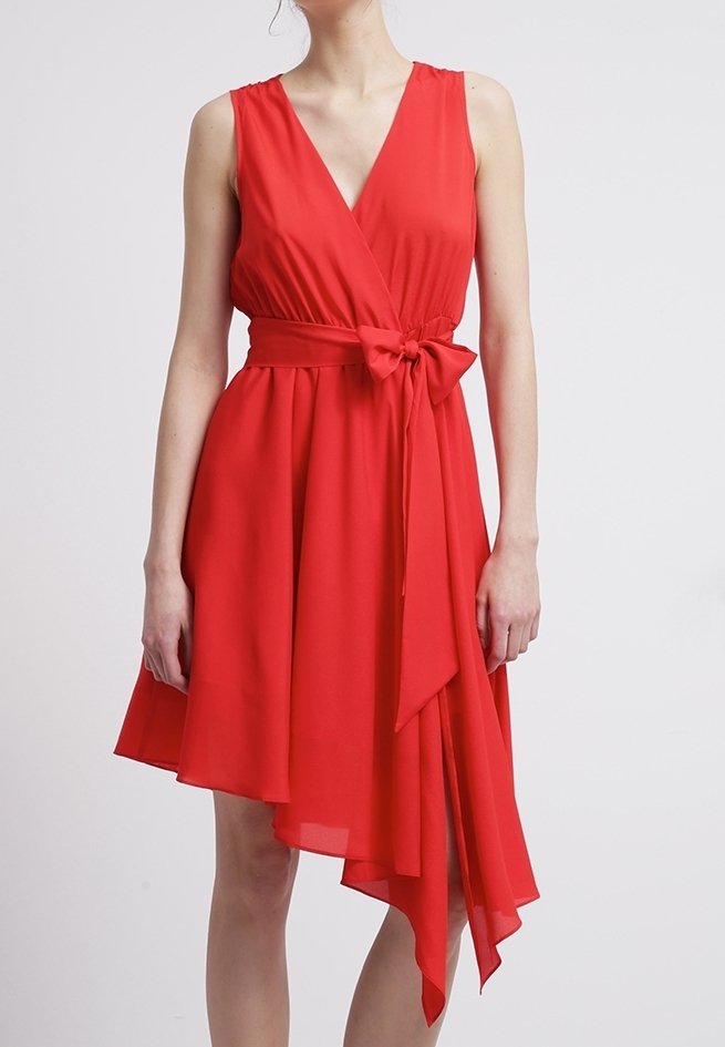 Robe rouge sans manches avec encolure en V, taille cintrée nouée en un nœud, et jupe asymétrique à volants portée par une personne debout.