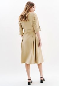 Abito khaki con cintura, maniche a tre quarti, pinces sul retro e lunghezza fino al polpaccio, abbinato a scarpe con tacco a blocco nere.