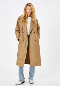 Abrigo trench de color marrón claro con un diseño de doble botonadura, bolsillos delanteros y hombreras. Llevado sobre una blusa blanca y jeans azules.