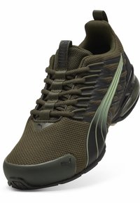 Zapatilla deportiva de malla verde con acentos negros y verdes, parte superior texturizada, suela flexible y un sistema de cordones destacado. Logo icónico en el costado.
