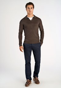 Man som bär en brun långärmad tröja över en vit skjorta, marinblå byxor och bruna läderskor, stående mot en enfärgad vit bakgrund.