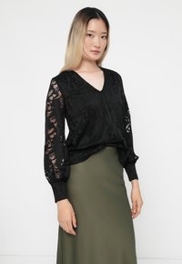 Μαύρη μπλούζα με διάφανα δαντελένια μανίκια, σχέδιο V-λαιμού, υφασμάτινη υφή, συνδυασμένη με εφαρμοστό ελαιόχρωμο φούστα.