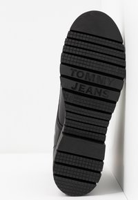 Semelle d'une botte noire avec des crampons profonds et « TOMMY JEANS » embossé sur la semelle extérieure en caoutchouc, taille 37 visible.