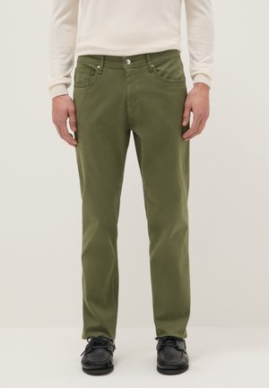 Homme portant un pantalon droit vert olive, des chaussures noires à lacets et un pull crème à manches longues, debout devant un fond uni.
