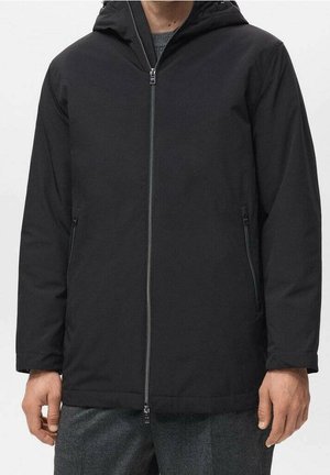 Mann trägt eine schwarze Kapuzenjacke mit Frontreißverschluss und seitlichen Reißverschlusstaschen, kombiniert mit grauen Hosen.