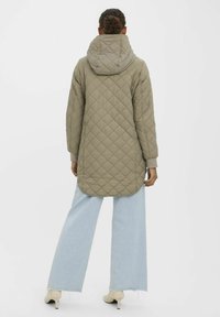 Veste longue en olive matelassée avec une capuche, présentant un ourlet arrondi et des poignets en côtes, associée à un jean large bleu clair.