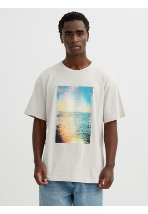 Hombre vistiendo una camiseta blanca con un estampado colorido de océano y atardecer, combinado con vaqueros de mezclilla azul claro, de pie frente a un fondo blanco liso.