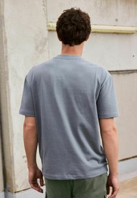 Tricou scurt, de culoare gri deschis, cu guler rotund, realizat dintr-un material moale. Spatele are un design curat, fără grafică sau modele.