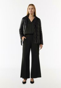 Schwarze gesteppte Daunenjacke mit breiten Revers, kombiniert mit einem passenden schwarzen Oberteil und schmal geschnittenen Hosen, abgerundet mit schwarzen High Heels.