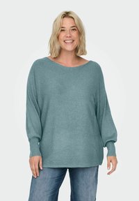 Mujer sonriente con cabello corto rubio que lleva un suéter de canalé suelto en color teal y jeans azules, de pie contra un fondo claro y liso.