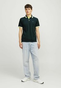 Polo shirt vert foncé avec des rayures jaunes contrastantes sur le col et les manches, associé à un jean en denim clair et des baskets blanches.