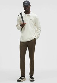 Kremowy golf z długim rękawem i kołnierzykiem, z haftowanym logo; zestawiony z oliwkowo-zielonymi spodniami slim fit i czarnymi sneakersami.