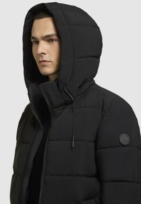Schwarze Daunenjacke mit Kapuze, ausgestattet mit gestepptem Design, leichtem Material und einem runden Logo auf der linken Schulter. Mit Reißverschluss vorne.