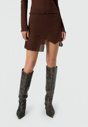 Kvinde iført en brun asymmetrisk mininederdel og langærmet top kombineret med mørke mønstrede knælange støvler.