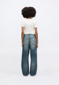 Φαρδιά τζιν παντελόνια σε ανοιχτό μπλε με τσέπες μπροστά και πίσω, και εμφανές ετικέτα μάρκας στο πίσω μέρος. Λευκό εφαρμοστό τοπ με κοντά μανίκια.