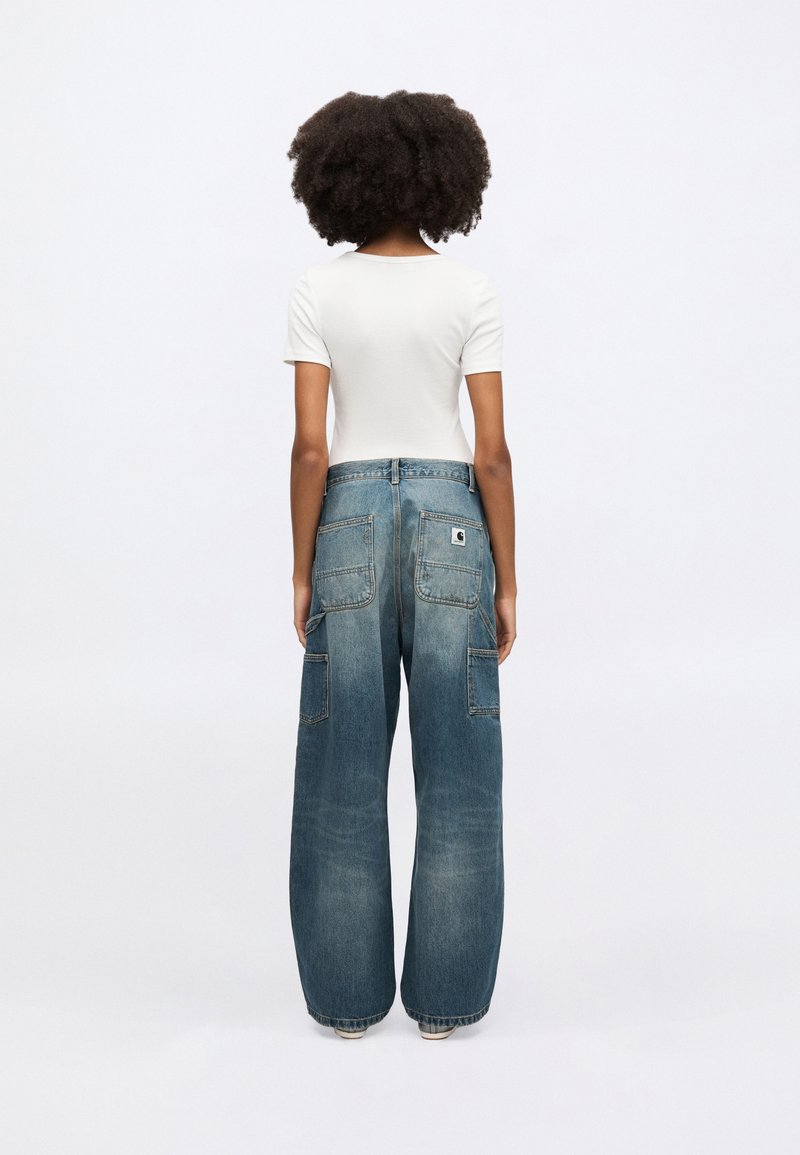 Vidbenede denimbukser i lyseblå med lommer foran og bagpå samt synligt mærke bagpå. Hvid tætsiddende top med korte ærmer.