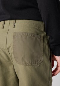 Pantalones verde oliva con un prominente bolsillo cuadrado. Presentan detalles de costura y una tela texturada, combinados con un suéter negro.