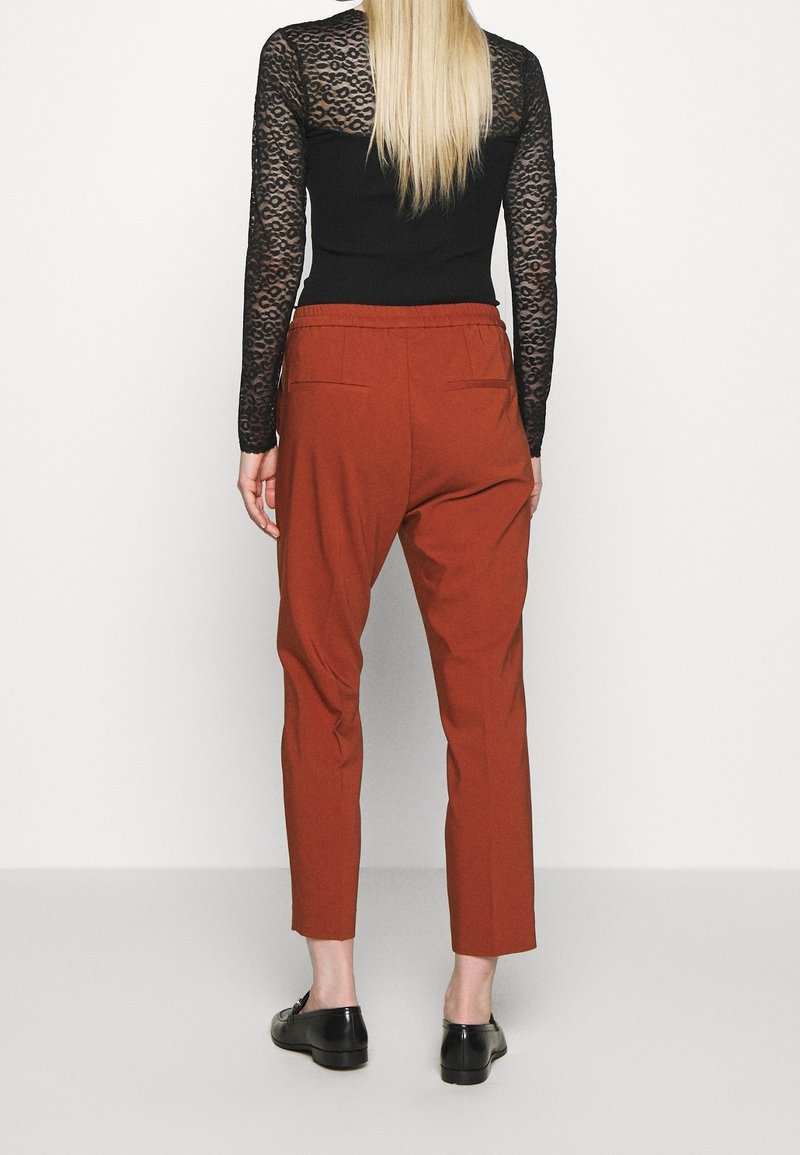 Femme aux longs cheveux blonds portant un haut noir transparent à manches longues et un pantalon court couleur rouille, debout devant un fond blanc uni.