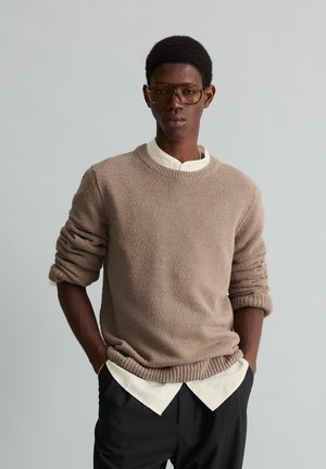 Giovane con capelli corti e ricci che indossa grandi occhiali marroni, maglione beige, camicia bianca e pantaloni neri, in piedi con le mani nelle tasche.
