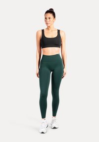 Schwarzer Sport-BH und dunkelgrüne Leggings aus dehnbarem Stoff. Die Leggings haben eine hohe Taille und eine glatte Textur. Das Modell trägt weiße Turnschuhe.