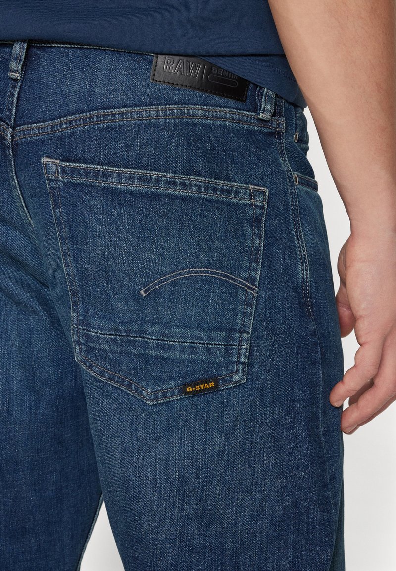 Jean en denim bleu foncé avec une poche arrière comportant une couture courbe et une étiquette jaune indiquant "G-STAR". Le matériau semble texturé.